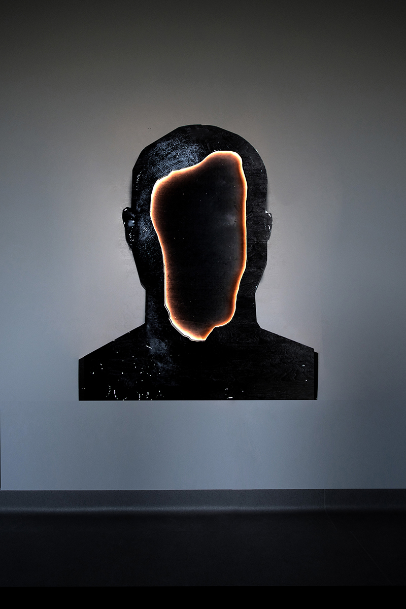 Portret mężczyzny pomalowany na czarno, w miejscu twarzy widnieje dziura, obramowana światłem