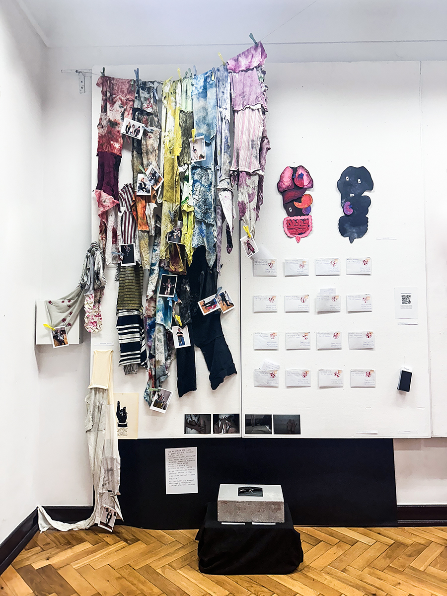 Wieloelementowa kompozycja prac na białej ścianie, składająca się z tkanin, listów i cegły