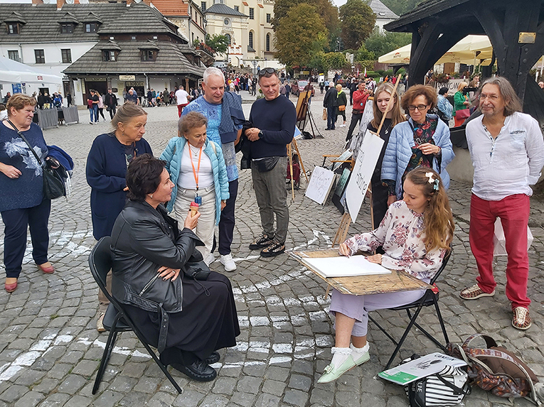 Dwie osoby na pierwszym planie, jedna pozuje, druga rysuje modelkę, a w tle publiczność przyglądająca się akcji. Całość dzieje się na rynku Kazimierza Dolnego.