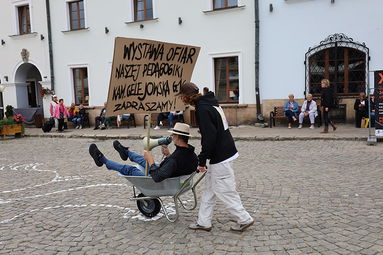 Dwie osoby w kadrze: jedna siedzi na tarce z megafonem i transparentem, druga pcha taczkę, wioząc postać po rynku Kazimierza Dolnego.