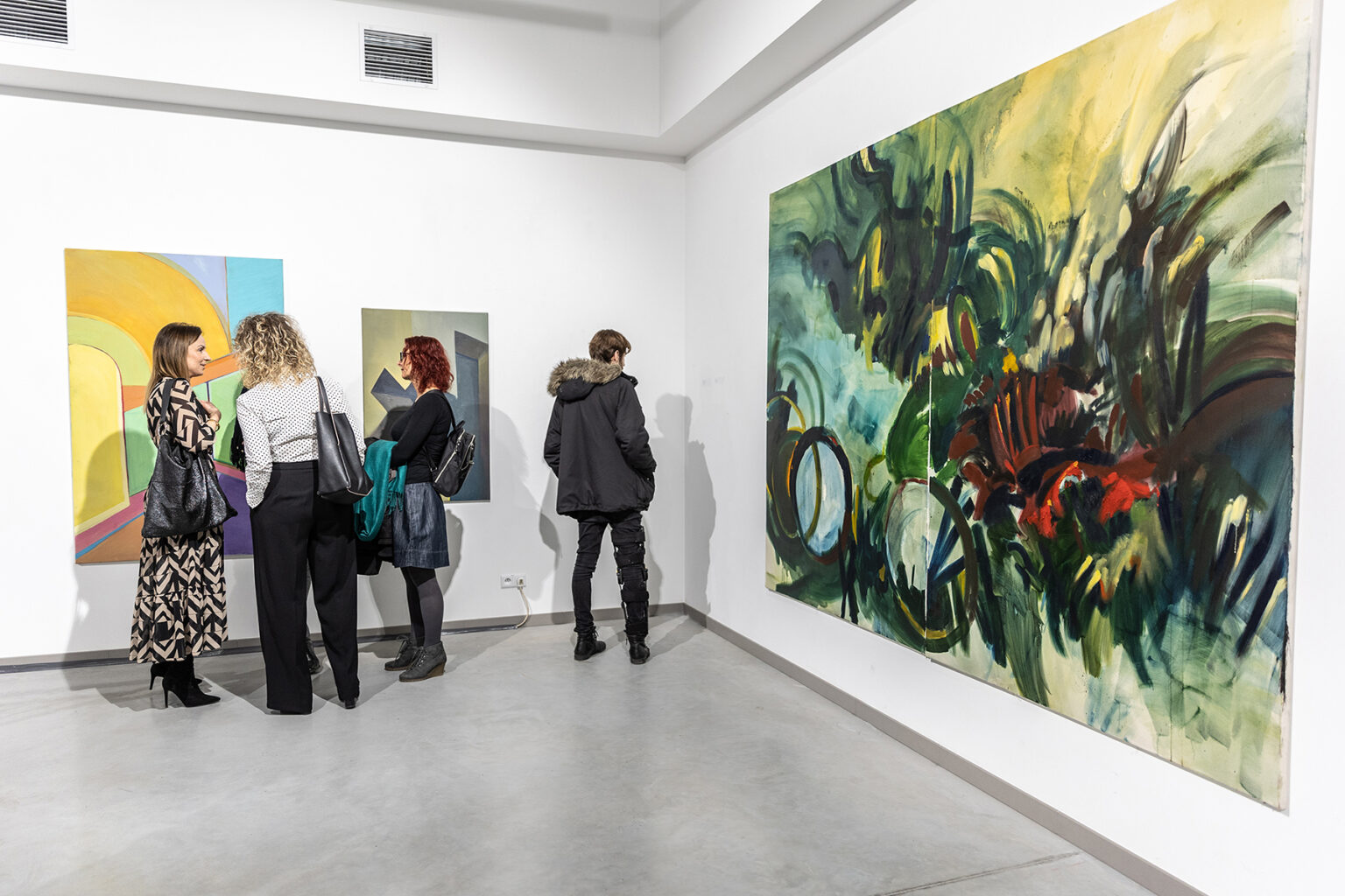 Widok wystawy w białym pomieszczeniu z ludźmi oglądającymi malarstwo o charakterze abstrakcyjnym