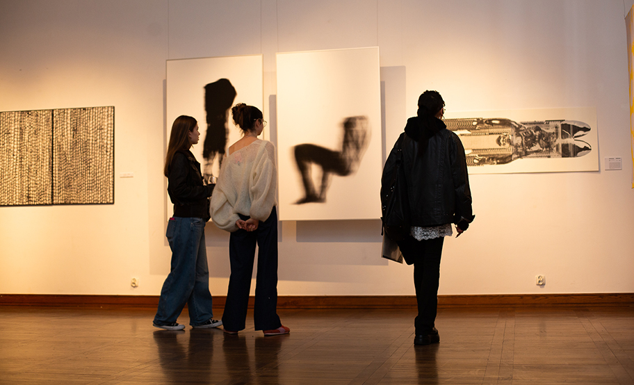 Trzy osoby oglądające grafiki na wystawie WG, na ścianie praca z ludzkimi sylwetkami