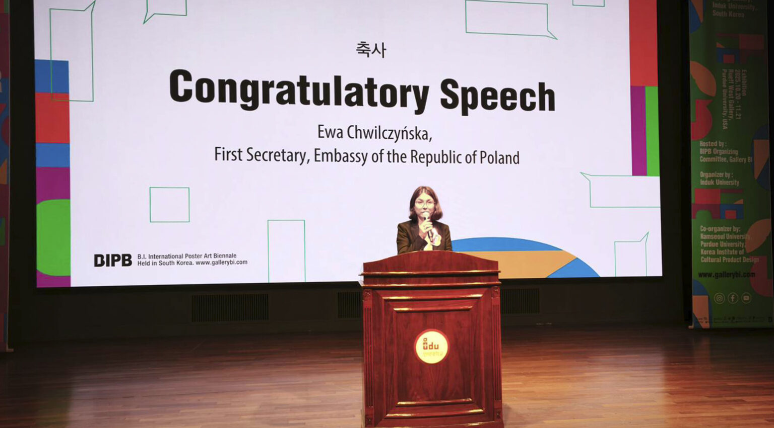 Mowa gratulacyjna dla zwycięzców podczas Biennale, wygłaszana na podium z prezentacją w tle.