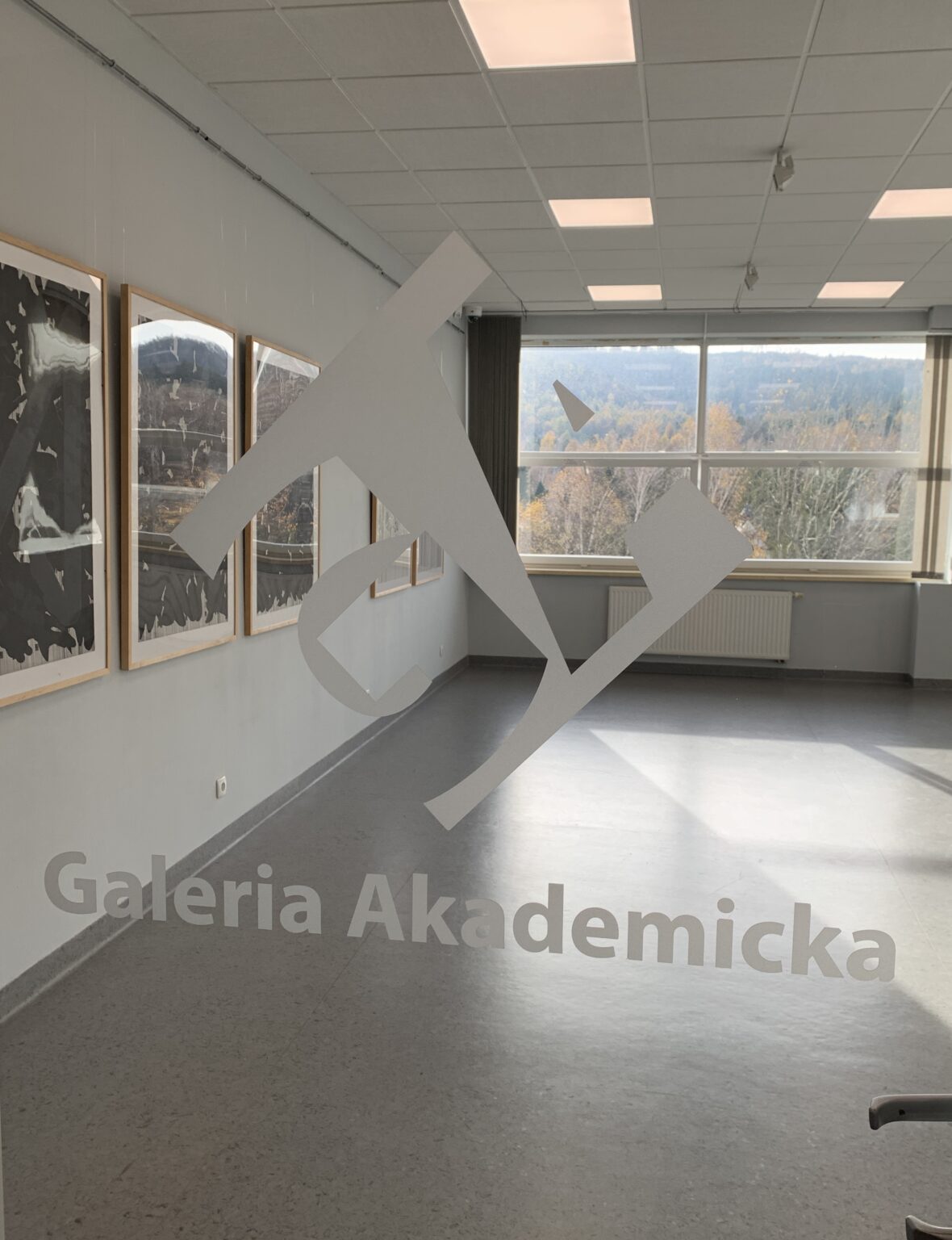 Widok wystawy przez szybę z logotypem Uniwersyteckiej Galerii Akademickiej.