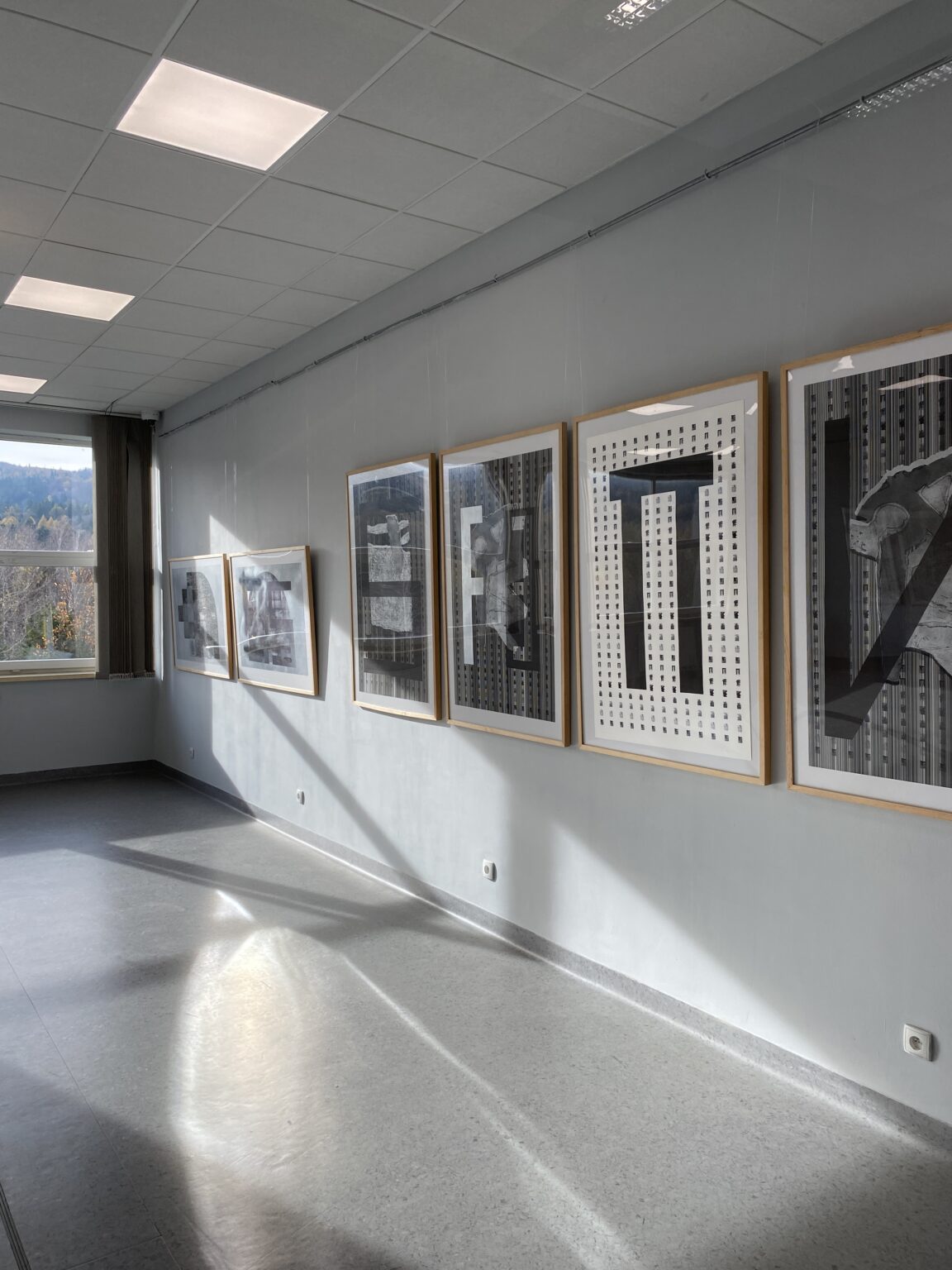 Widok 6 prac graficznych wiszących na szarej ścianie.