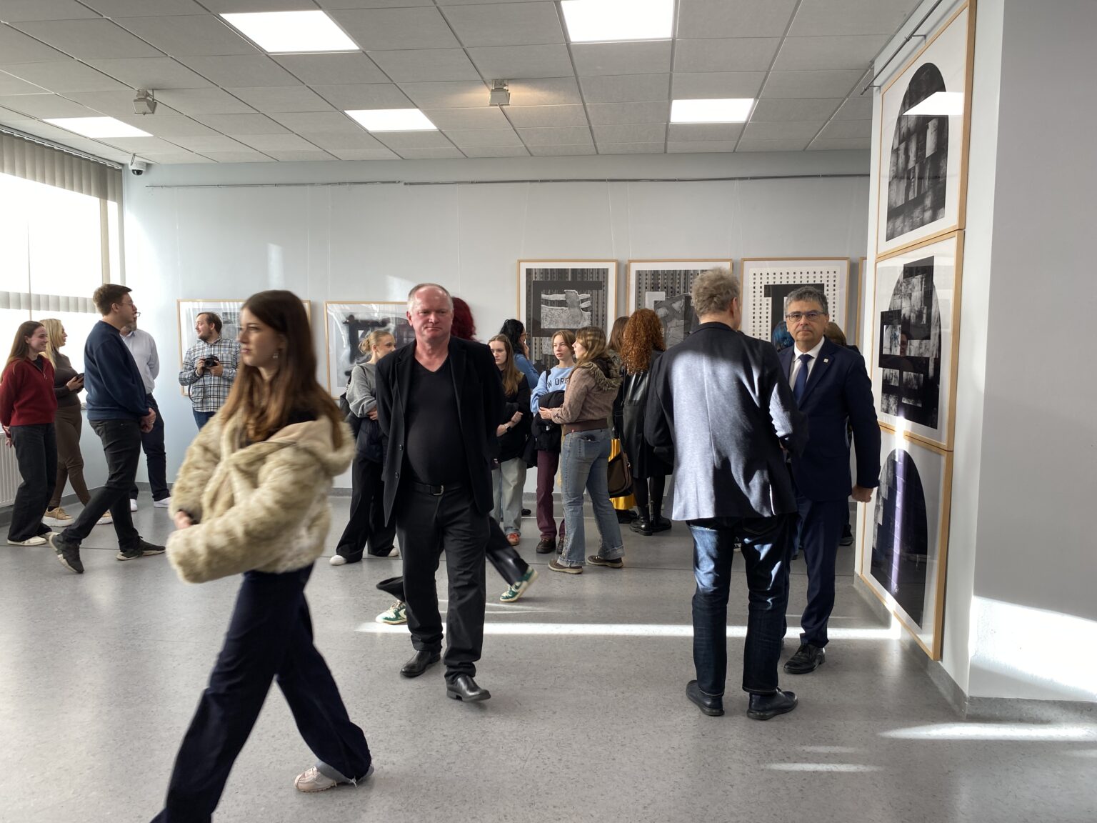 Widok otwarcia wystawy prac graficznych z osobami zwiedzającymi w sali.