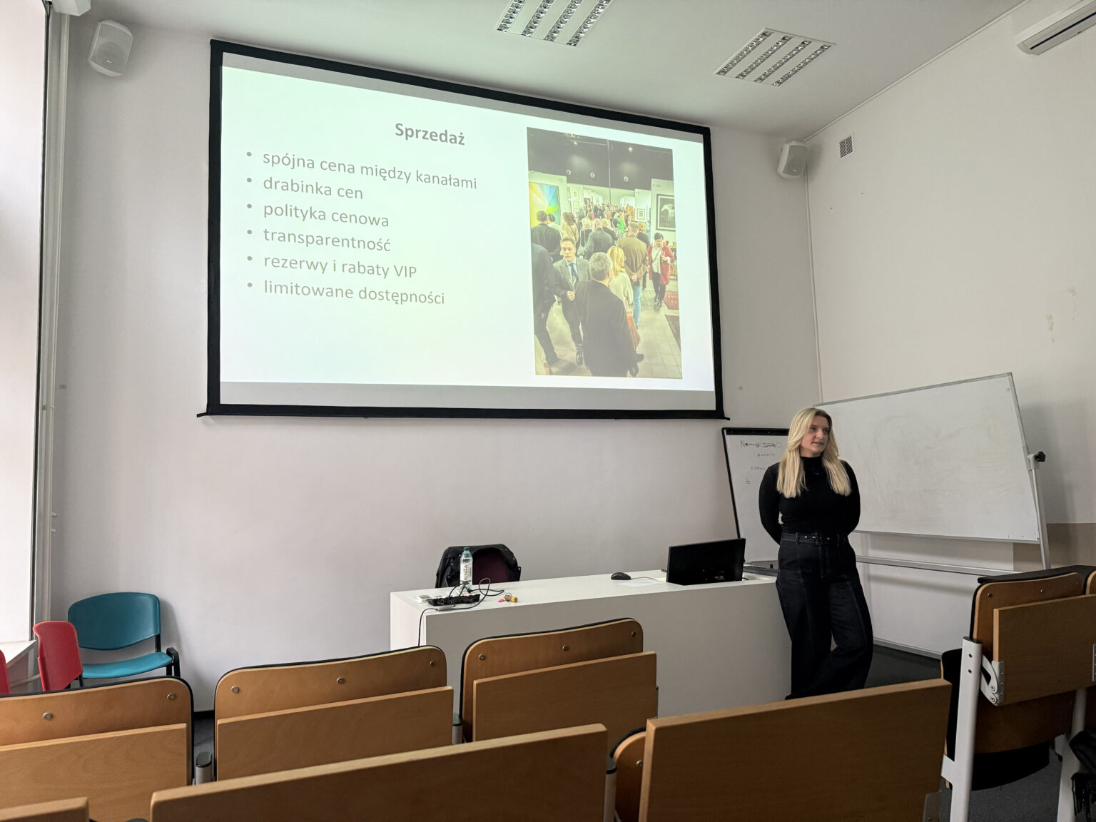 Prowadząca Katarzyna Marciniak w sali wykładowej na tle białego biurka i ekranu z prezentacją
