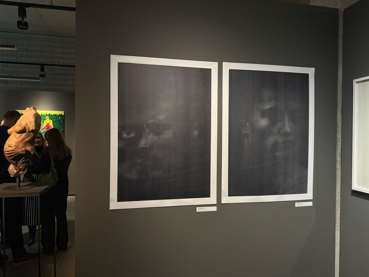 Widok dwóch grafik z twarzami na wystawie, prezentowanych na szarej ścianie w galerii.